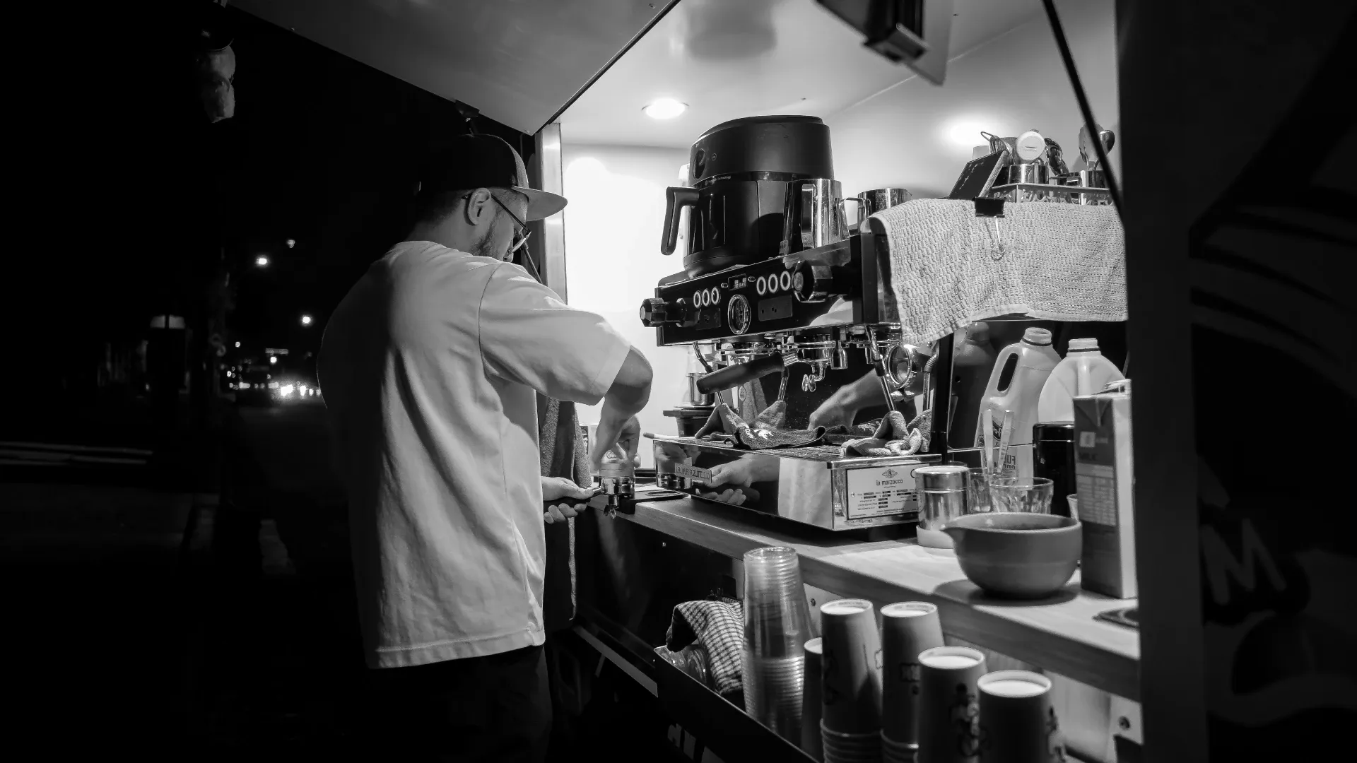 A person in a white t-shirt stands at a coffee truck espresso machine at night.
