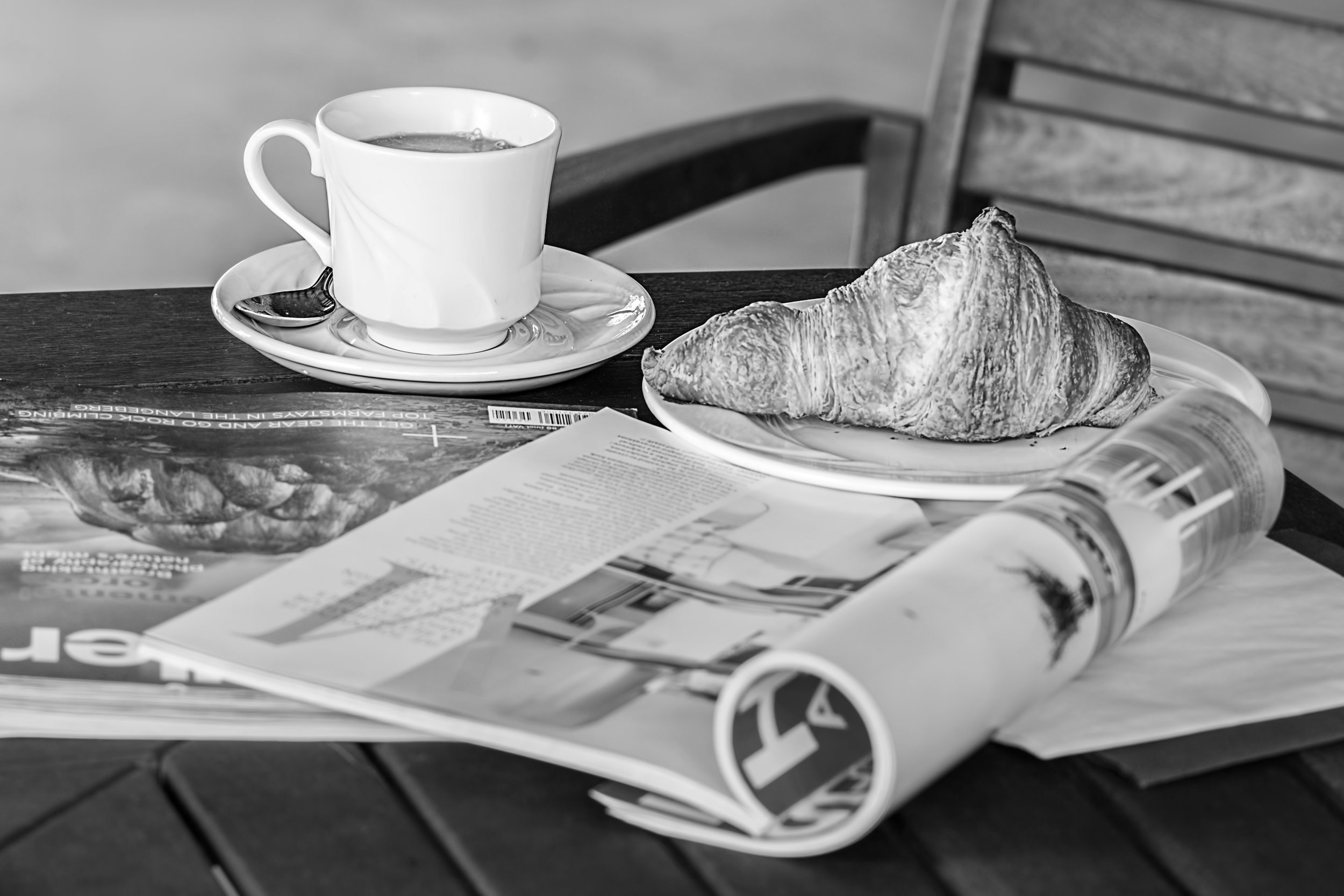 A coffee and a croissant on an open magazine atop a wooden table