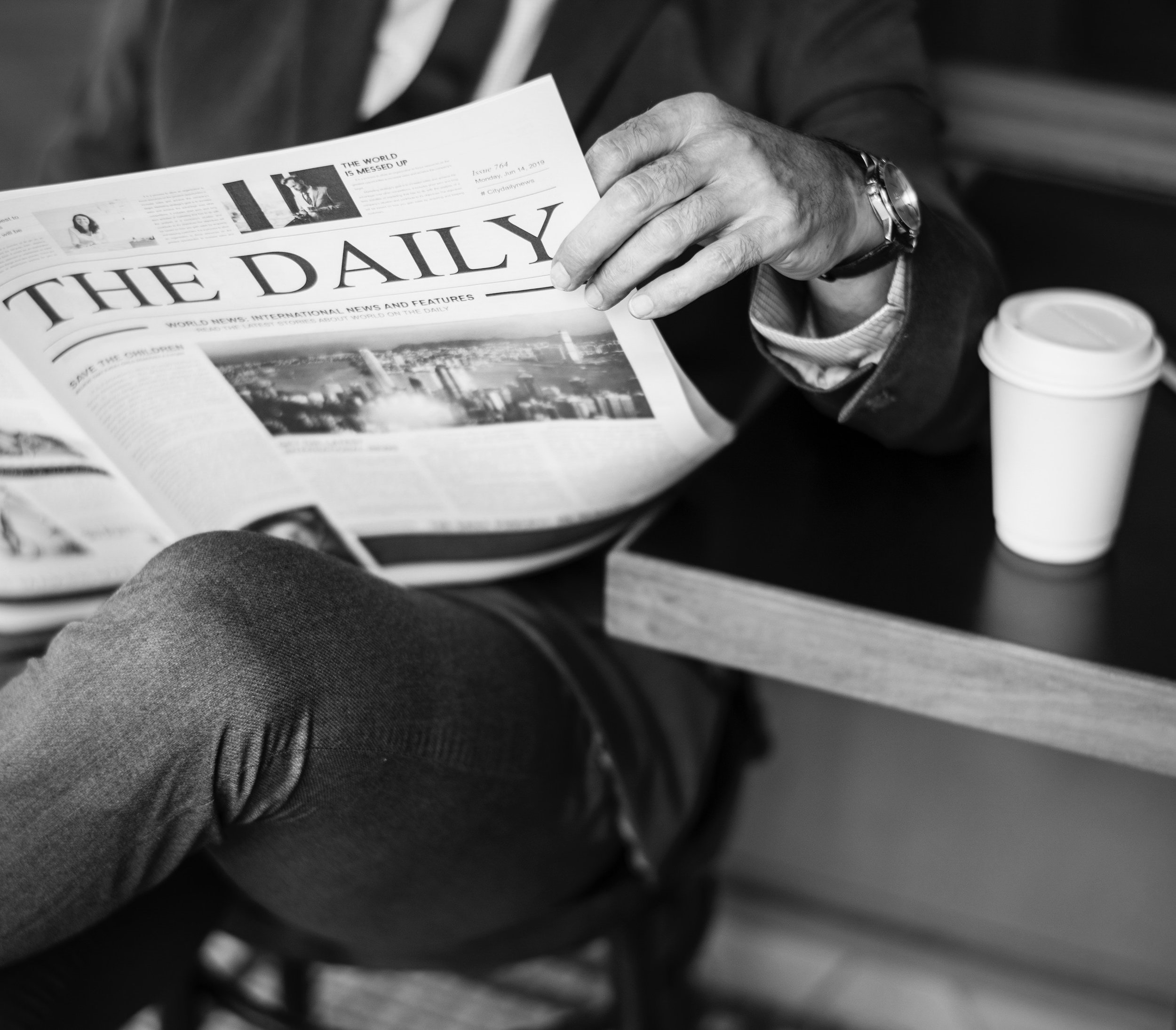 A man on a bench reads a newspaper with a paper coffee cup on a table beside him
