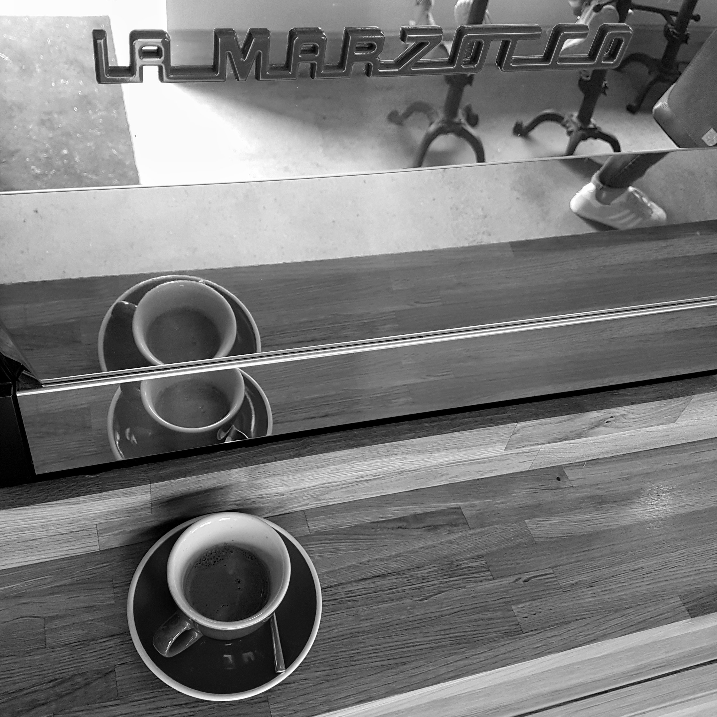 A shot of espresso sits on a counter in front of a shiny La Marzocco espresso machine