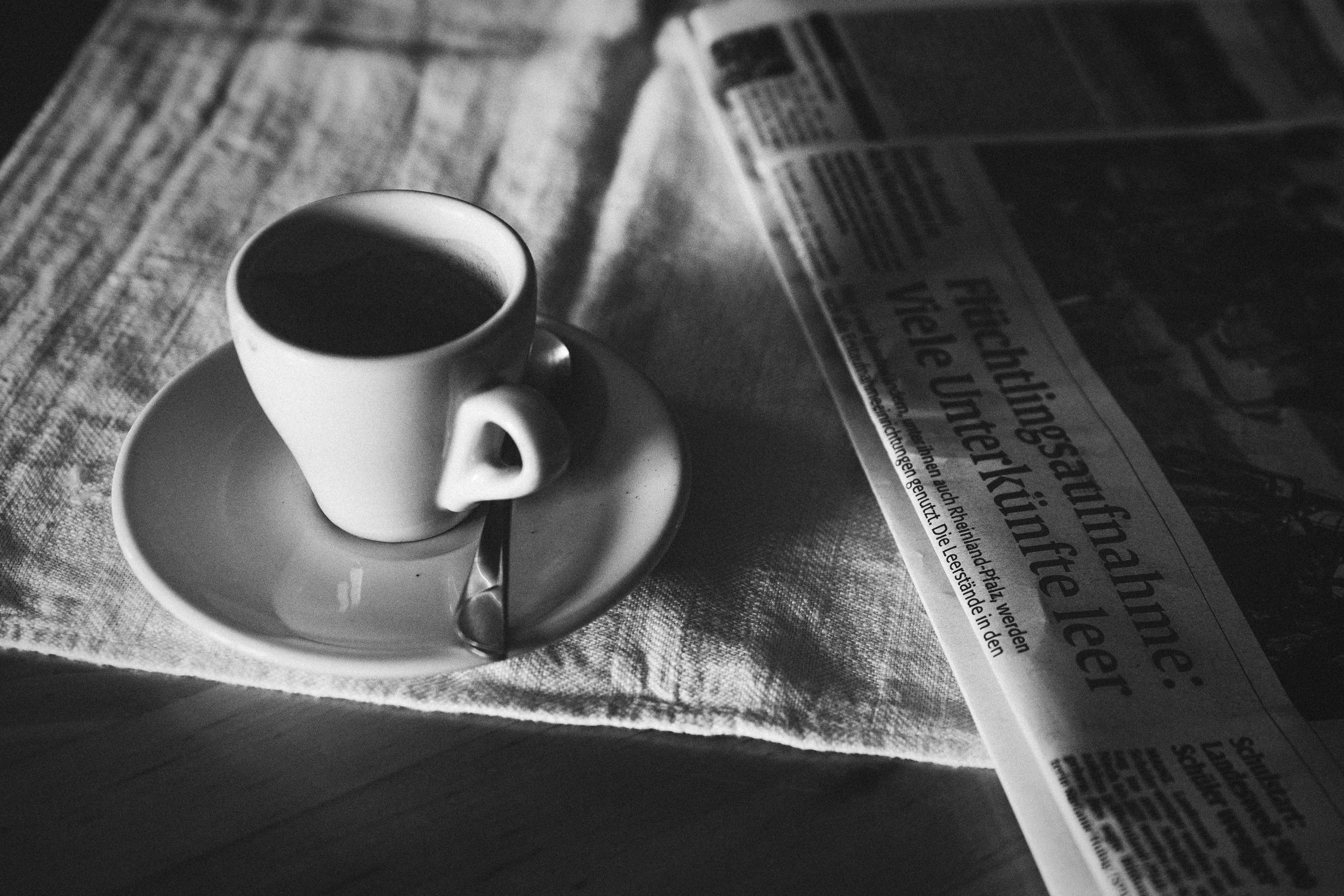 An espresso sits on a tablecloth beside a newspaper
