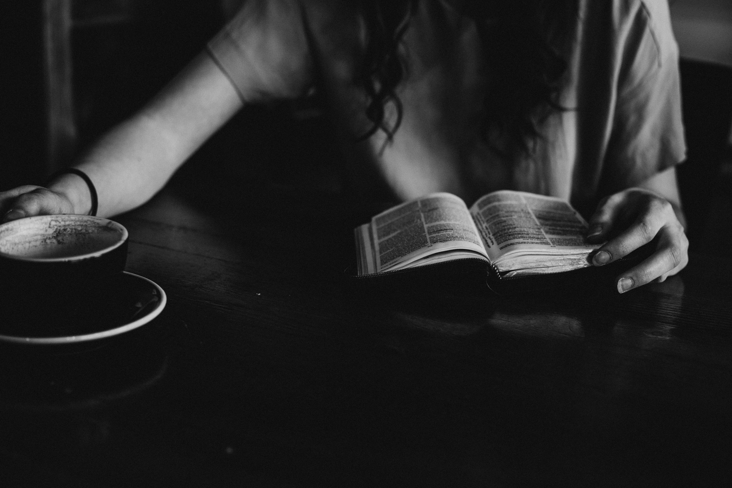 A woman reads a book at a table while reaching for a coffee