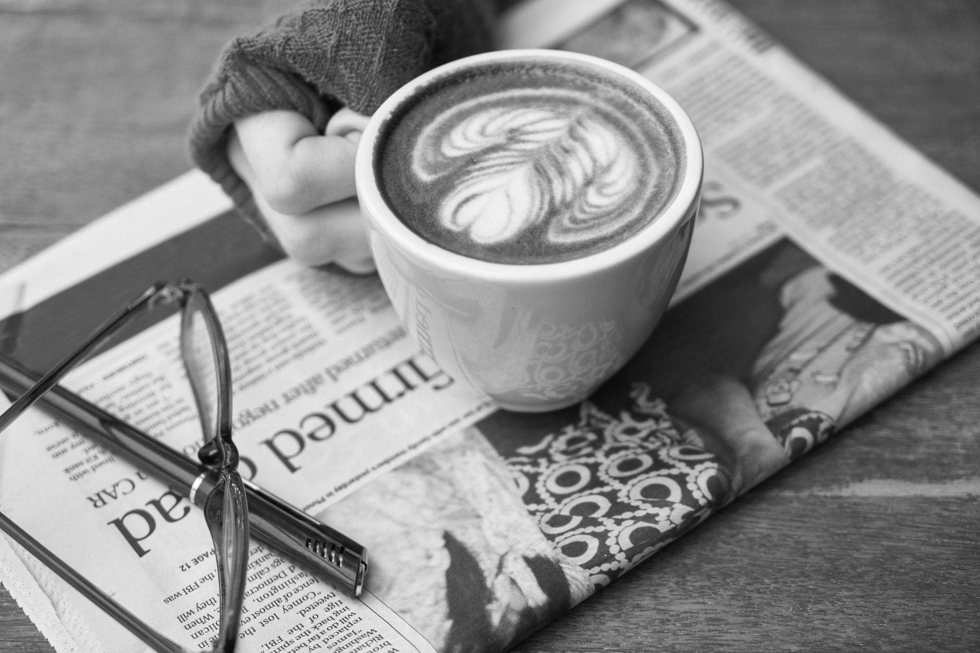 A hand holds a latte resting on a newspaper on a table.