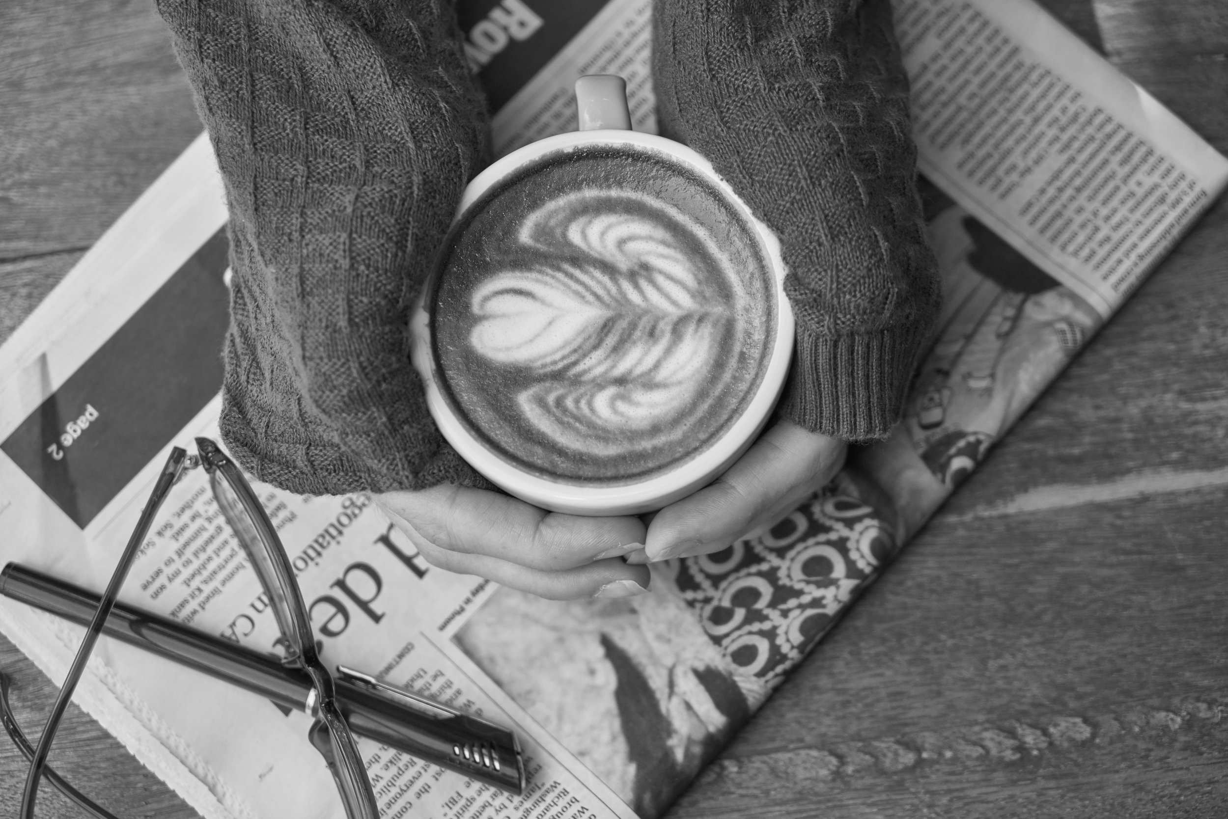 Two hands cup a latte, resting on a newspaper on a table.