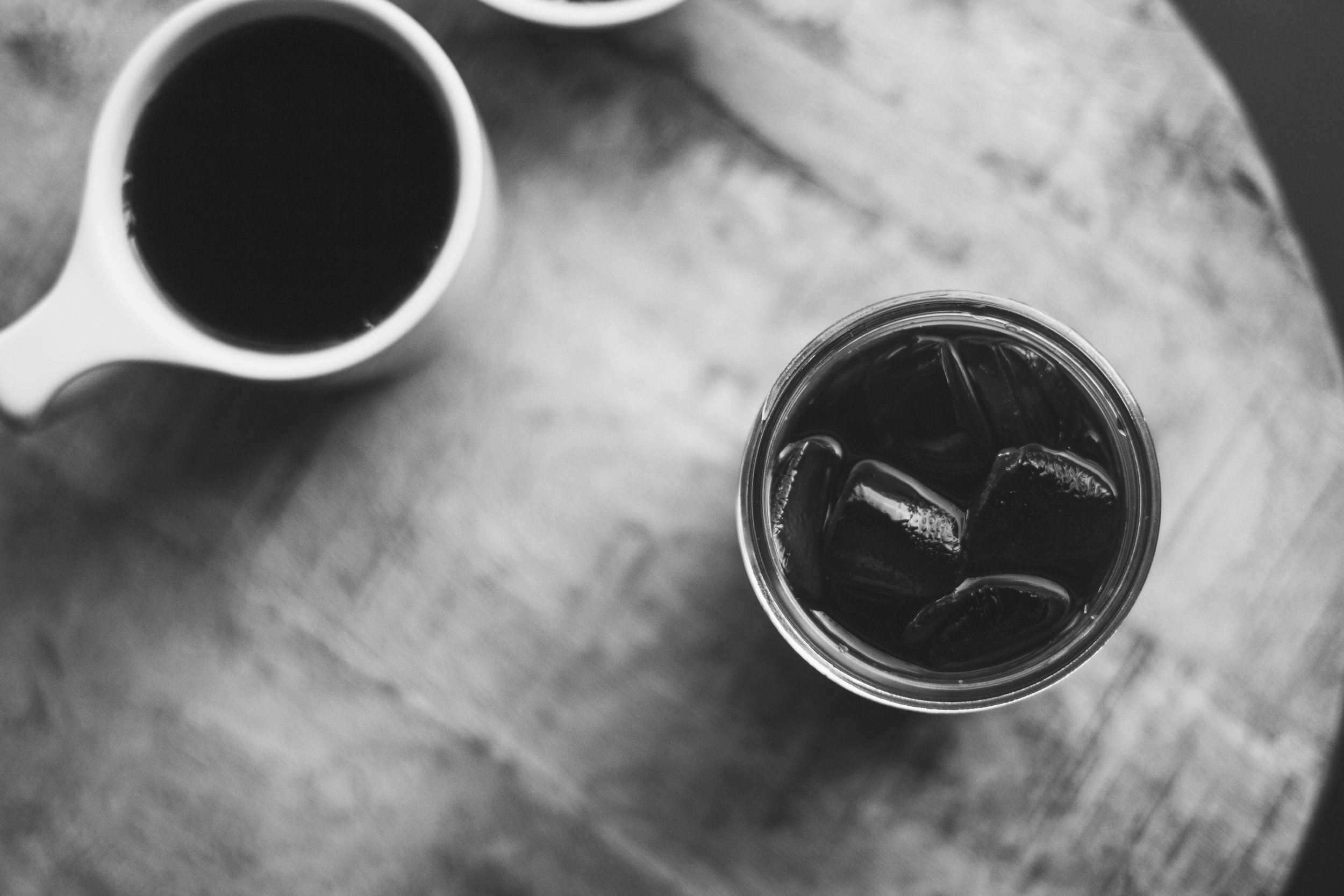 A cup of black coffee and a glass of iced coffee sit on a table, seen from above.