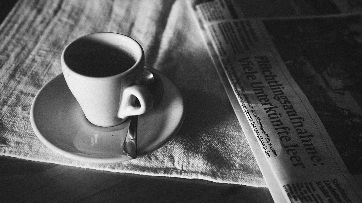 An espresso sits on a table next to a folded up newspaper.