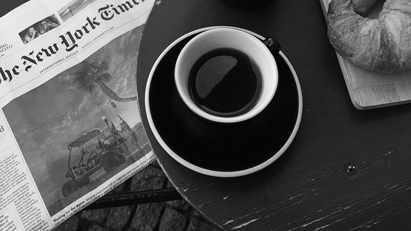An espresso sits on a wooden table next to a pastry and a newspaper