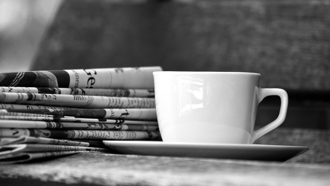 A cup of coffee sits on a table next to a stack of newspapers