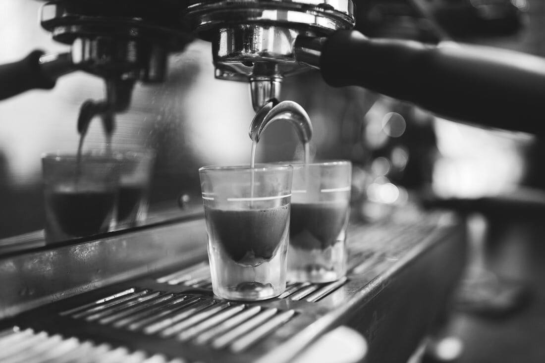 A shot of espresso pours into two glass cups sitting under a portafilter on a metal espresso machine tray