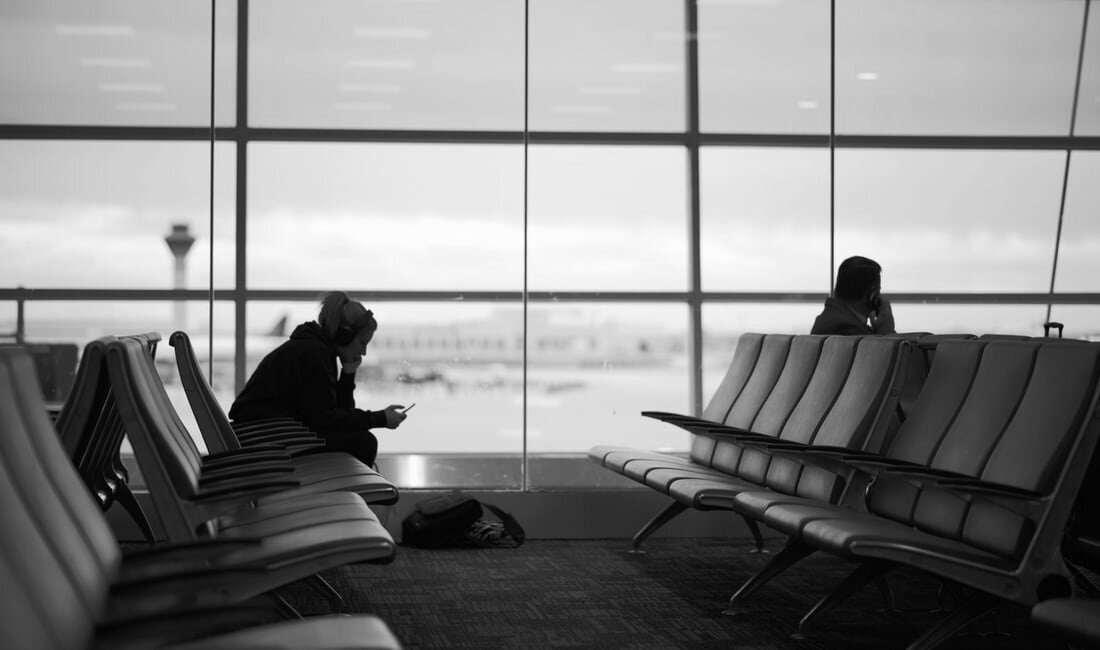 A person at the airport checks their phone while sitting on a chair with a glass window behind.