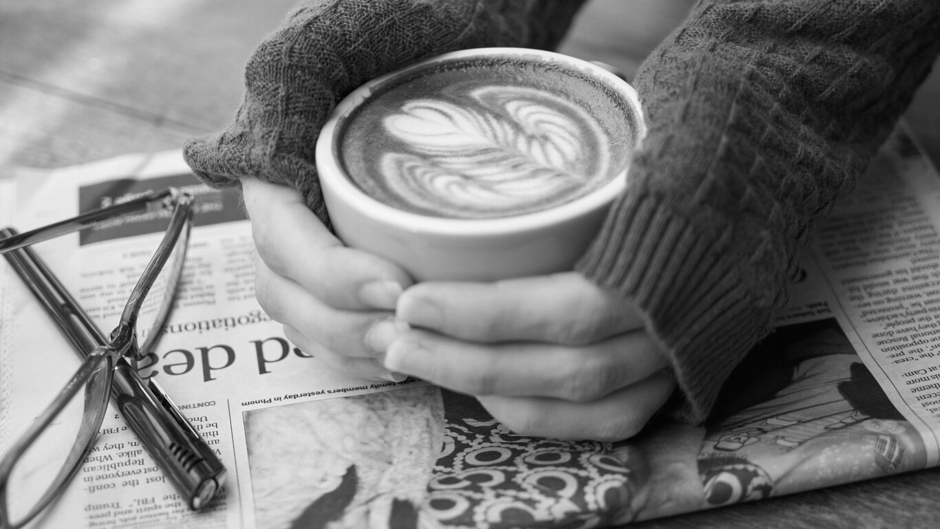 Two hands cupping a coffee cup with latte art, resting atop a newspaper