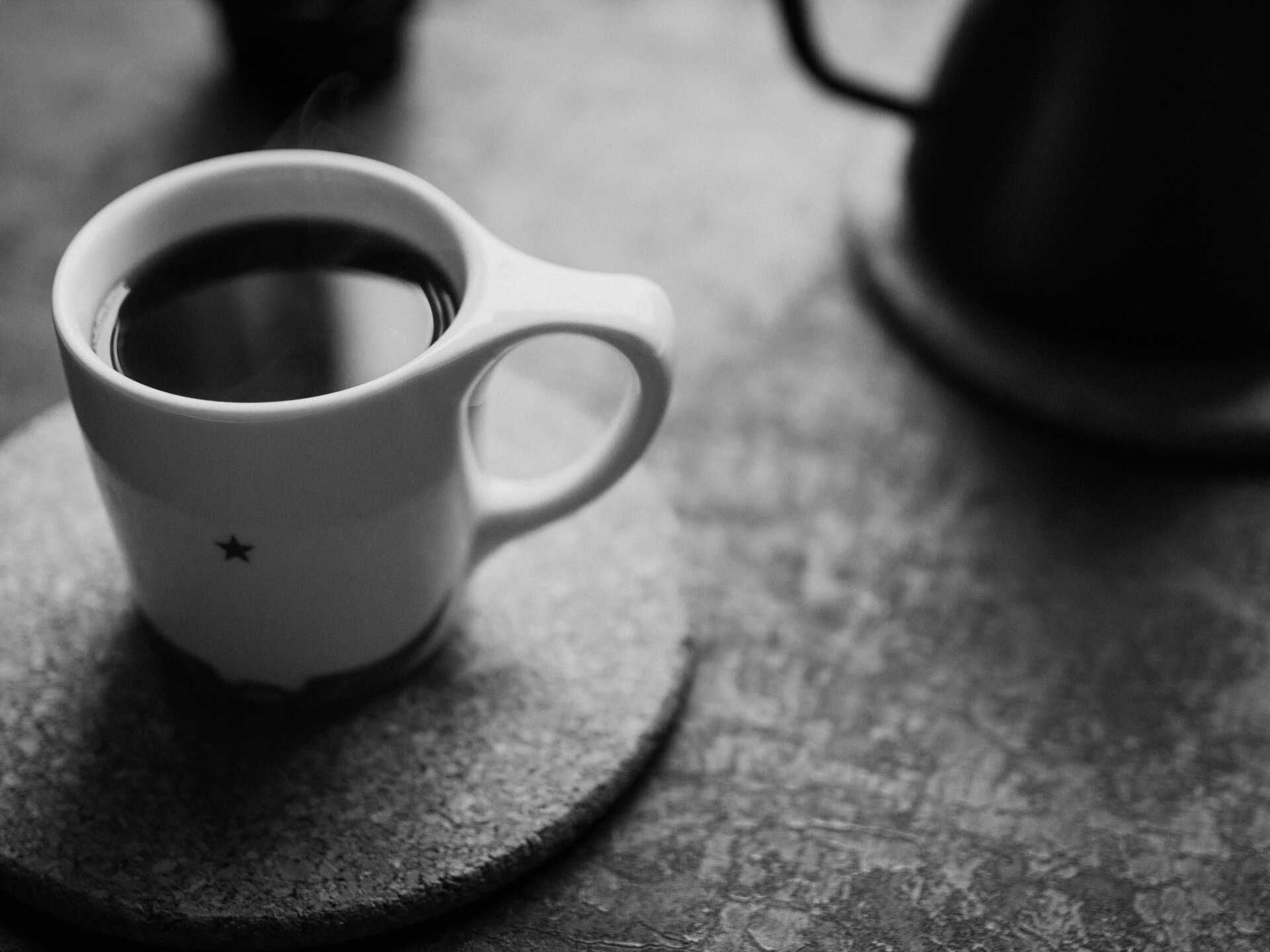 A white cup of black coffee sits on a cork mat on a table next to a kettle