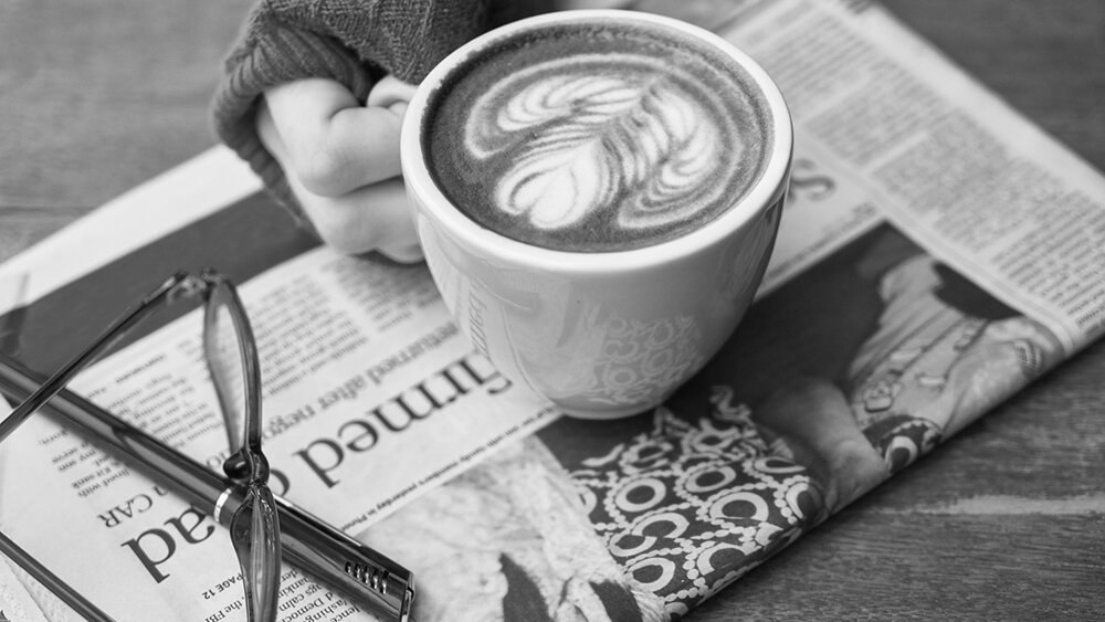 A hand holds a cup of coffee with latte art resting atop a folded newspaper