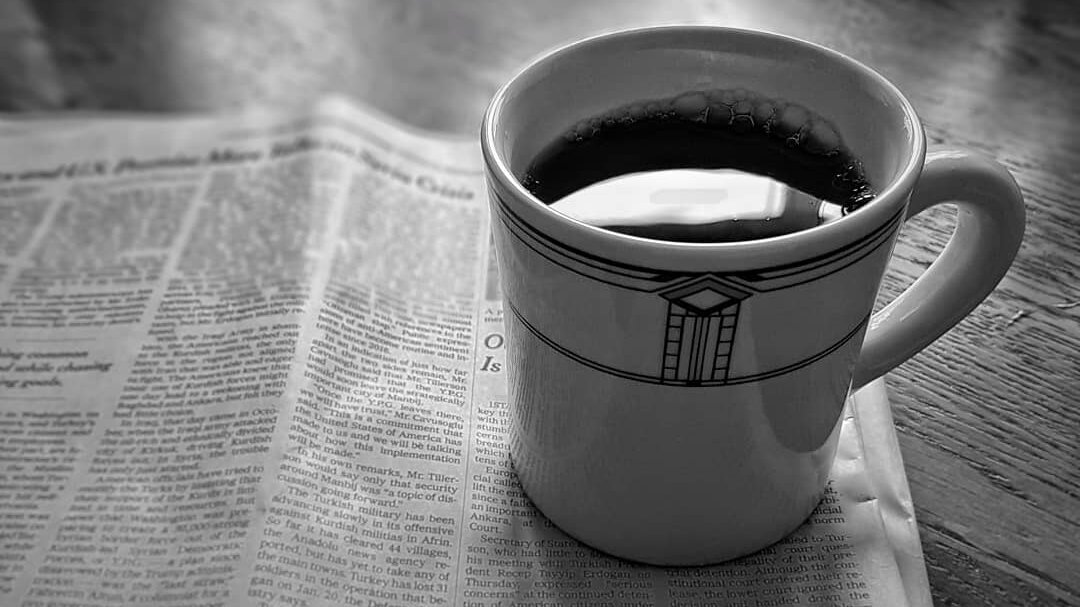 A diner mug reflects light from a window as it sits atop a folded newspaper on a table