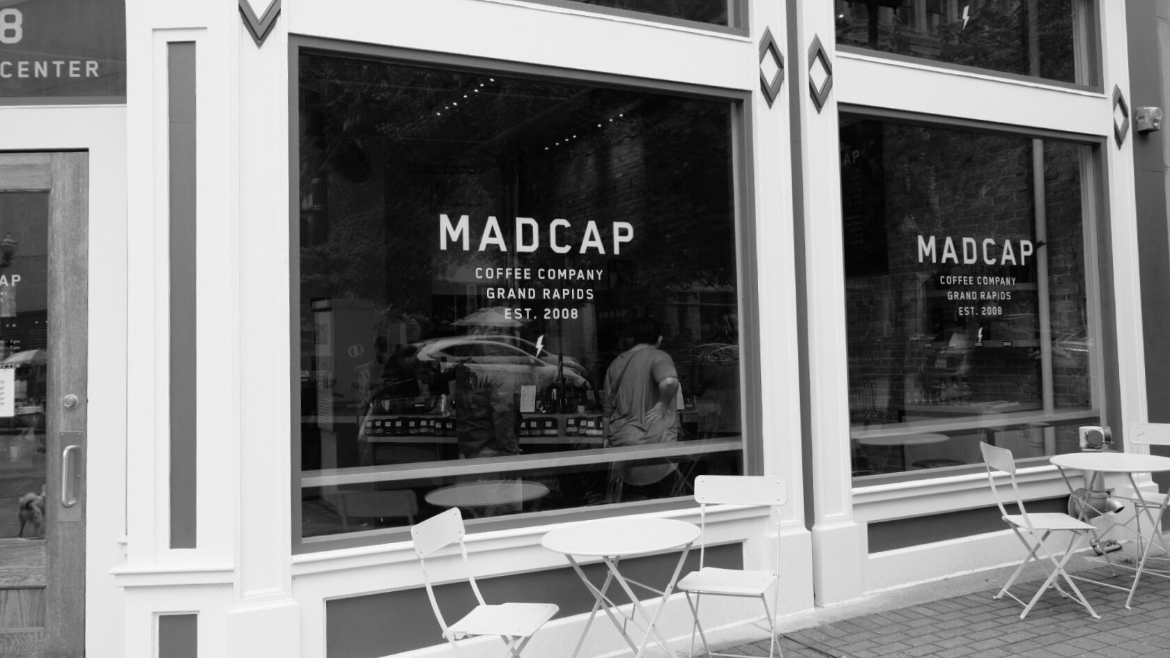 The exterior of Madcap Coffee in Grand Rapids, Michigan, showing glass windows with their logo and white outdoor tables and chairs in front
