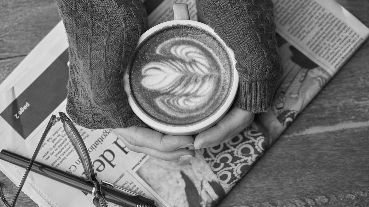 Two hands holding a coffee cup with latte art resting on a folded newspaper, seen from above