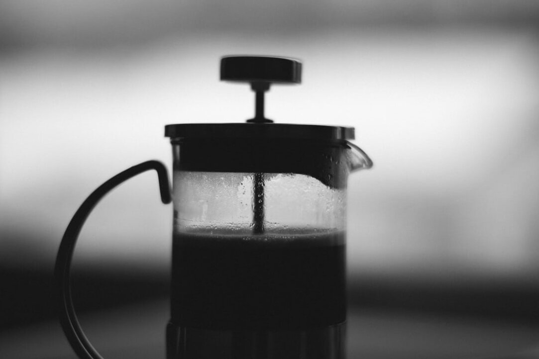 A French press is silhouetted against a light background. Via Unsplash