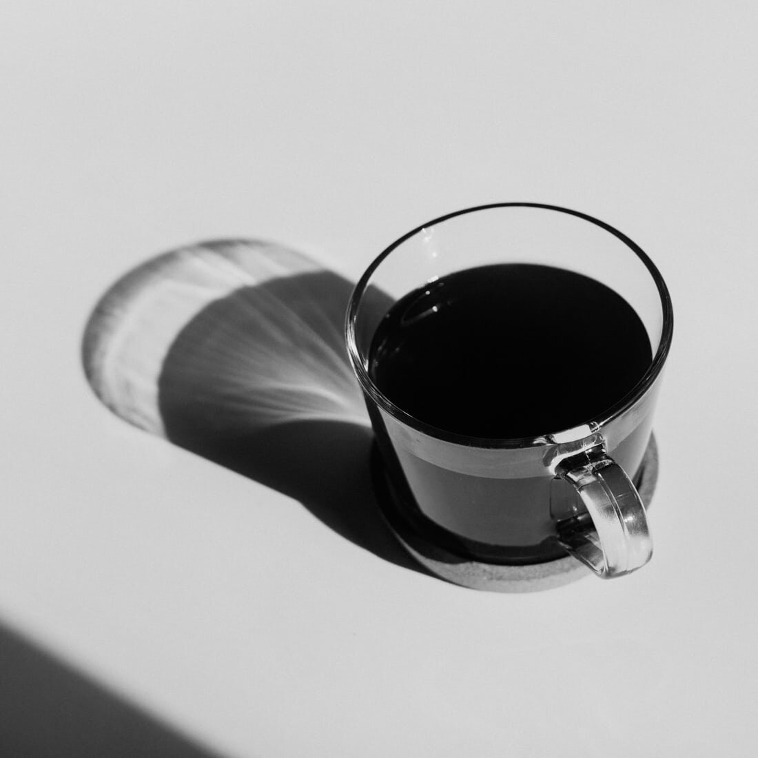A glass cup of black coffee sits on a table in the sunshine. Via Unsplash.
