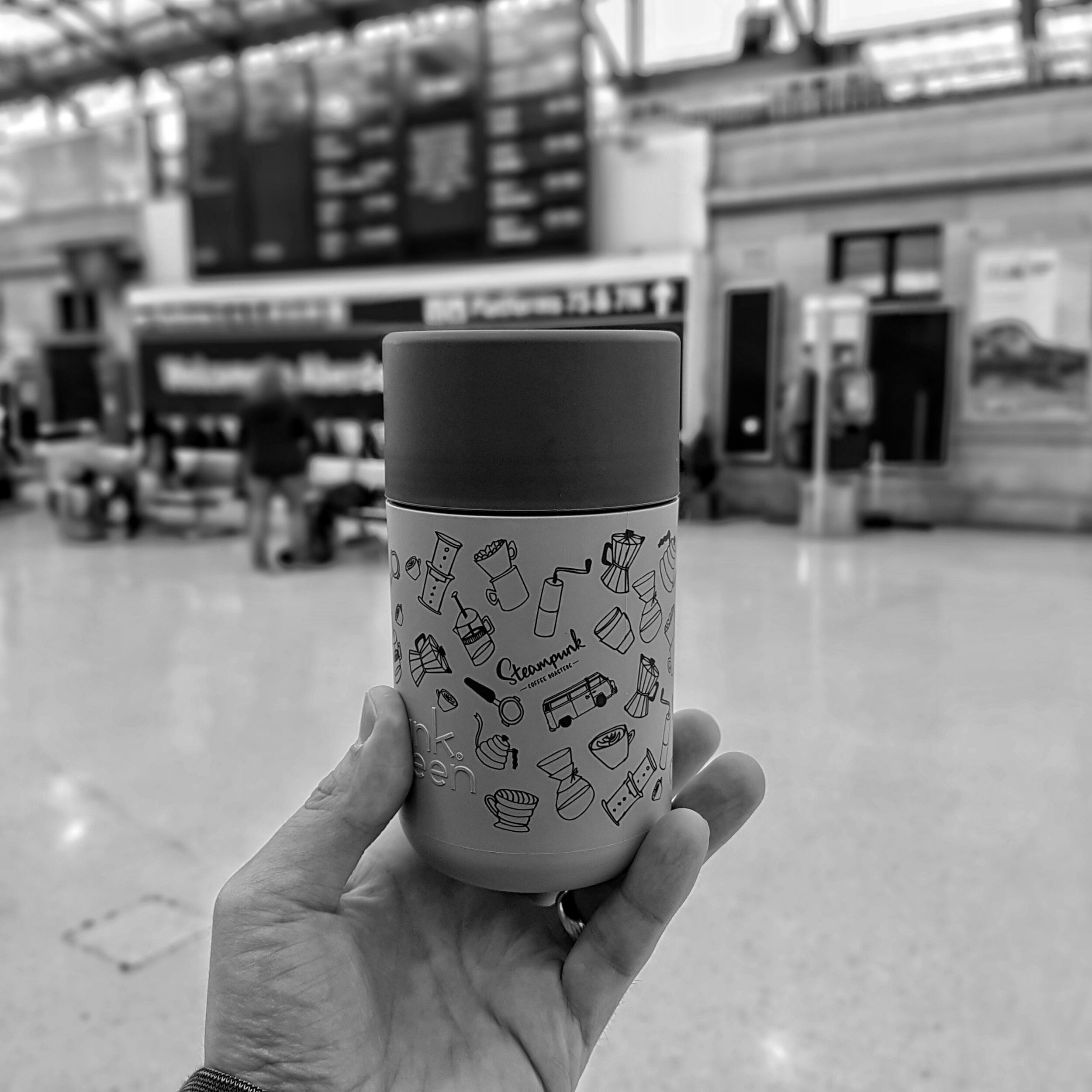 A hand holds up a reusable travel mug with a train station behind.
