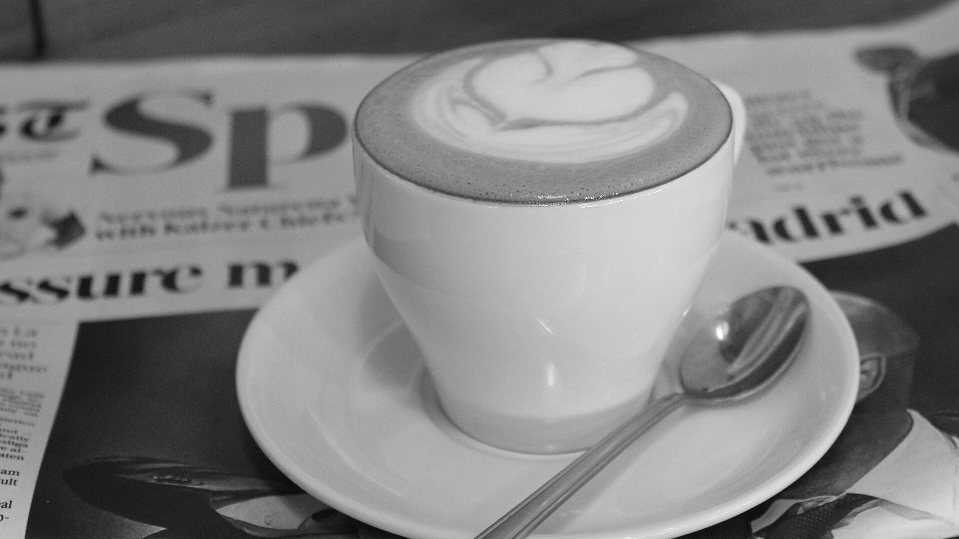 A white coffee cup with latte art and spoon sits atop a newspaper. Via Pixabay.