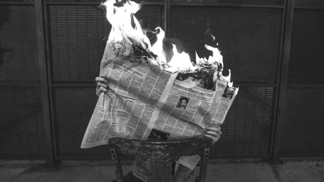 A person sits on a chair reading a newspaper which is on fire