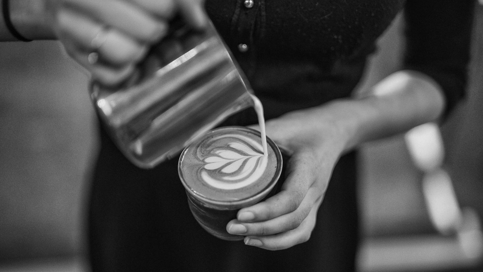 Close up of a barista pouring latte art with a jug into a cup, seen from above.
