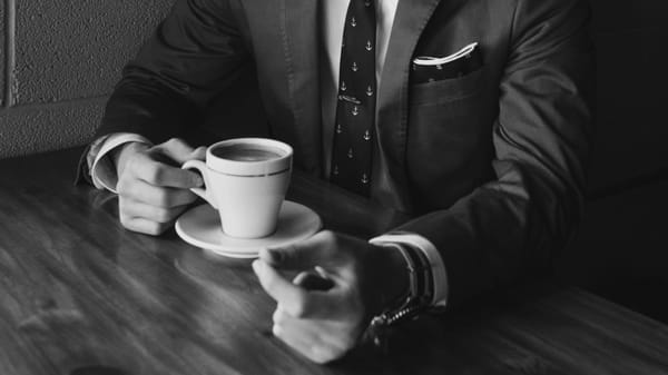 Close up of a person from the neck down wearing a suit sitting at a table with a cup of coffee in front of them. ra